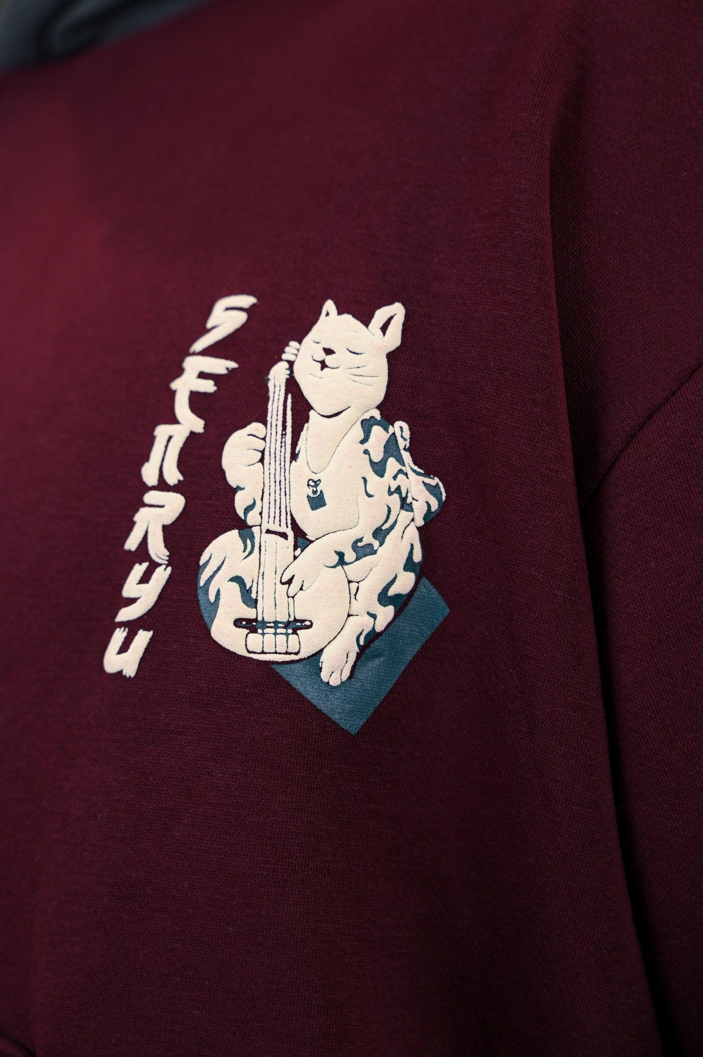 maroon hoodie with a contrasting dark grey hood features a cat strumming the shamisen